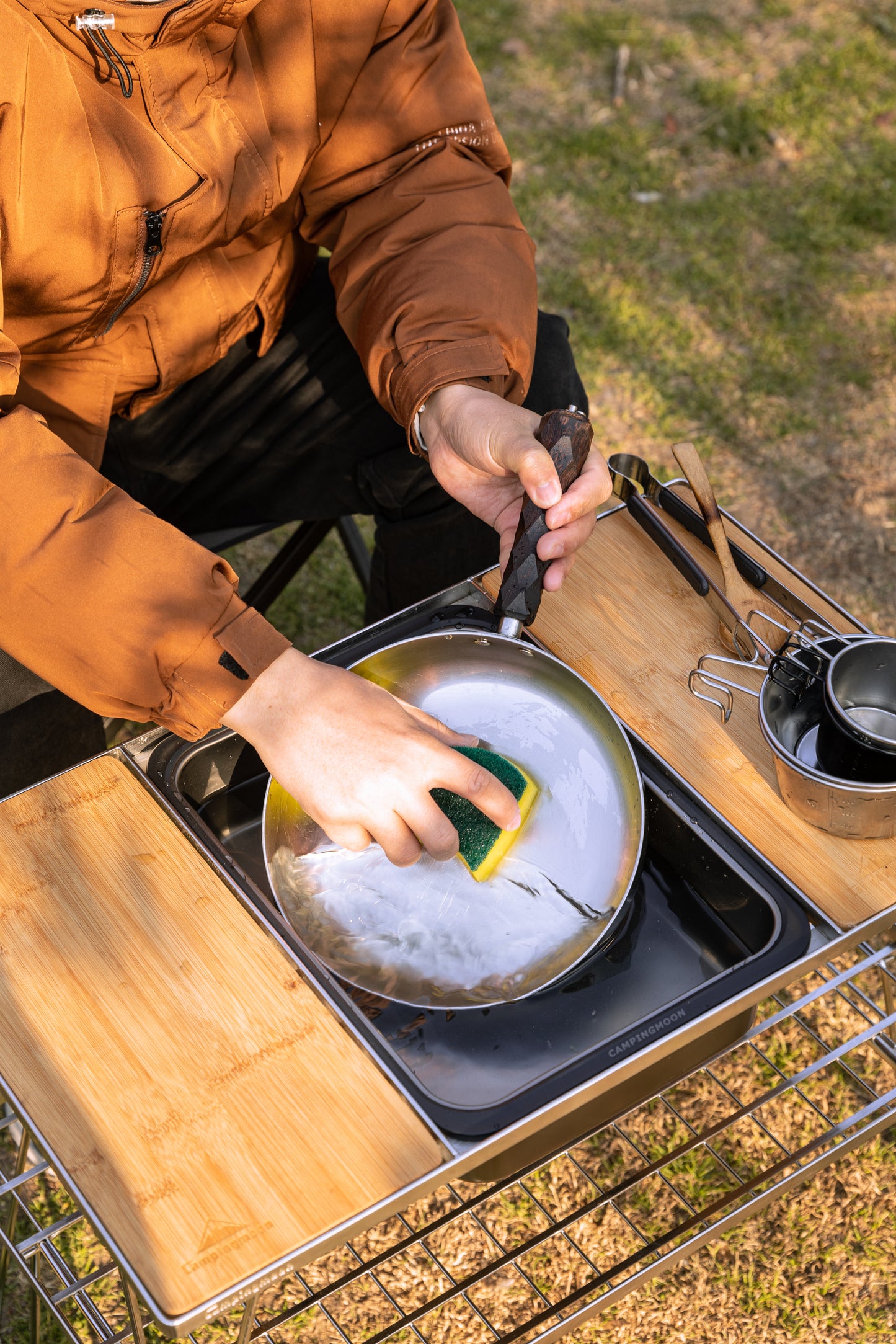 Person washing a pan in an outdoor setting using Nomadia CampStation Kitchen Sink on a modular camping table and grass in the background.
