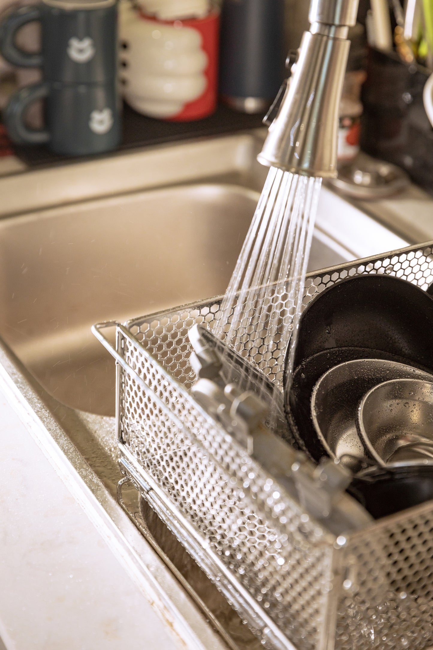 metal mesh basket used as dish rack with dishes being rinsed under a running faucet in a kitchen sink - multi-use camping gear - multi-purpose