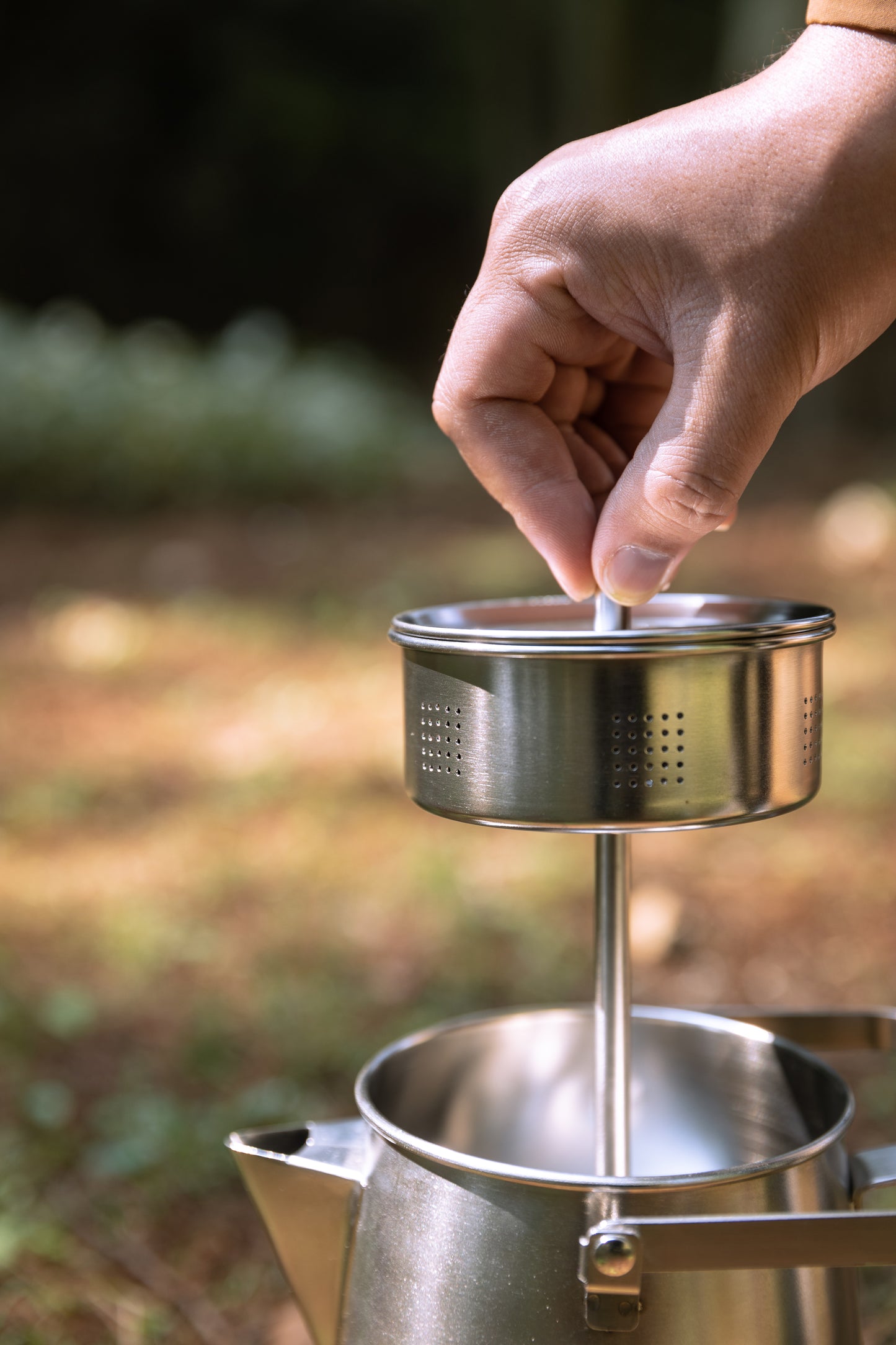 Hand using a metal tea strainer or coffee strainer over a pot outdoors Nomadia CampStation Stainless Steel Coffee Pot - outdoor coffee maker - glamping