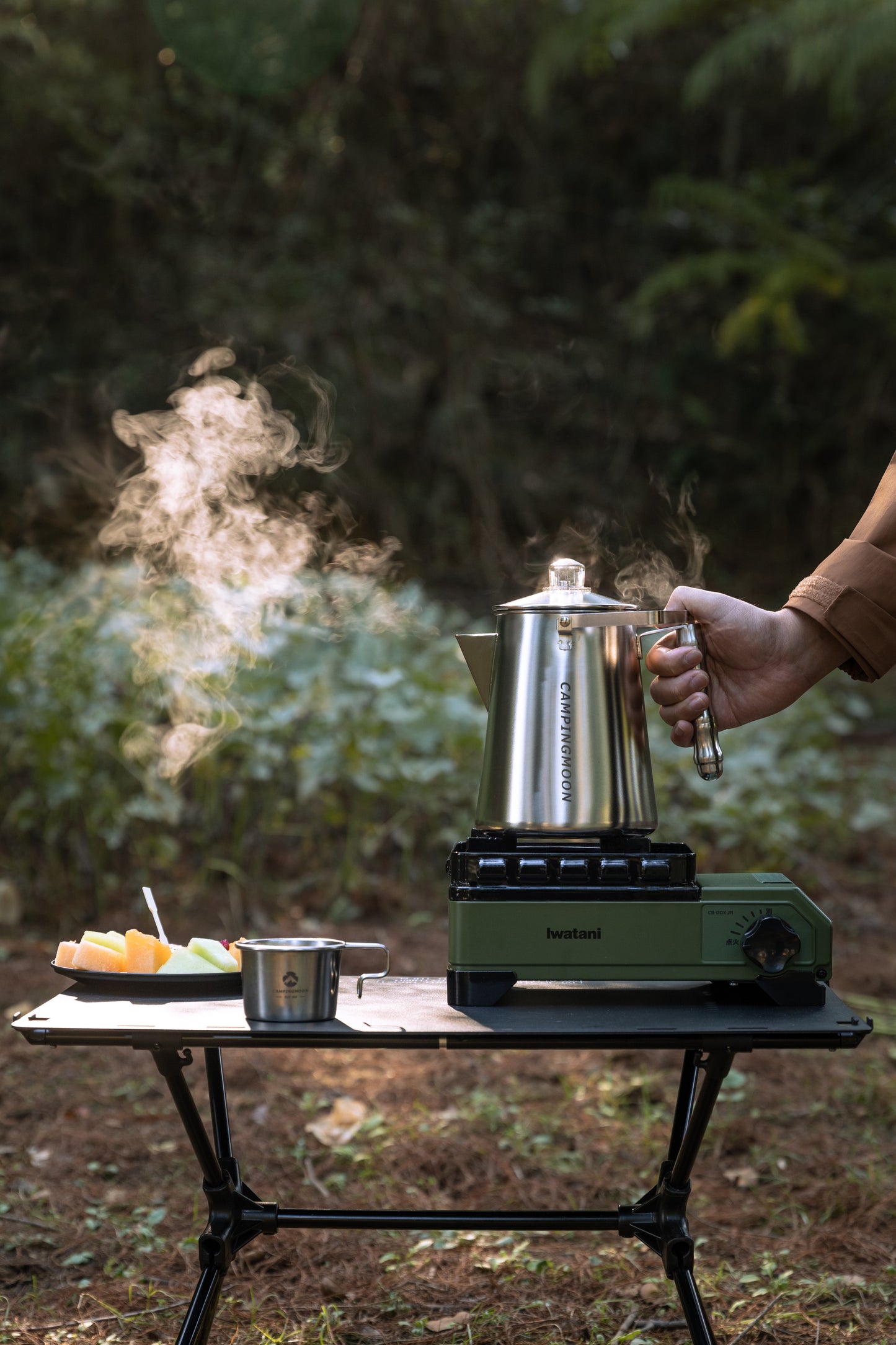 Person using a camping stove with Nomadia CampStation Stainless Steel Coffee Pot outdoors boiling coffee with steam - glamping