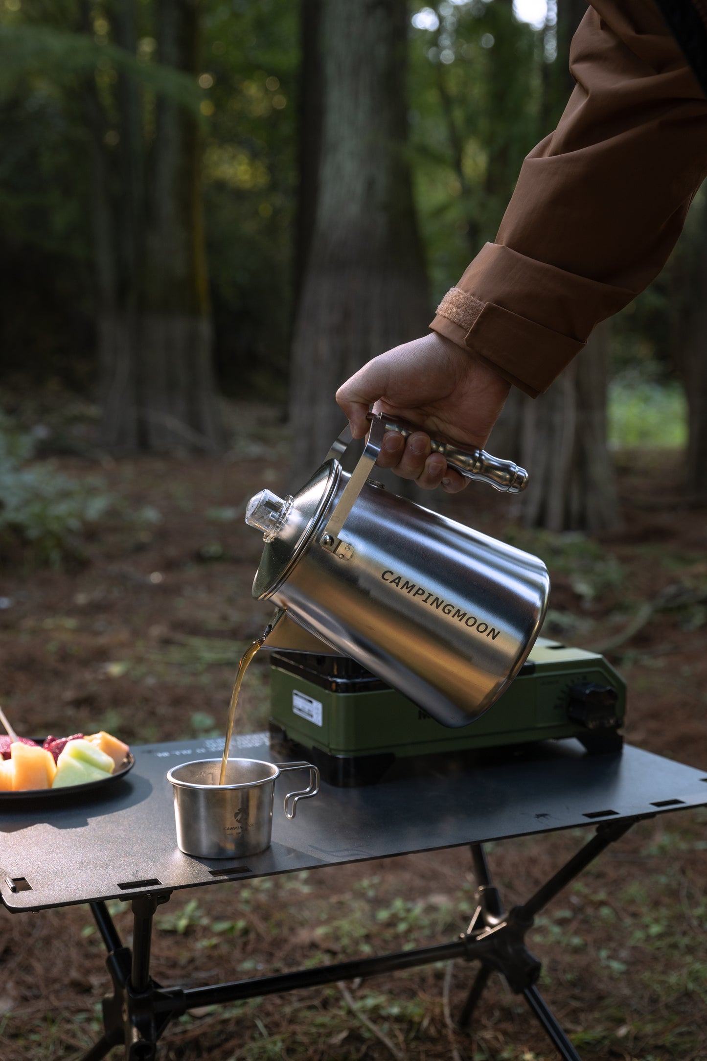 Person pouring hot coffee from a Nomadia CampStation Stainless Steel Coffee Pot into a cup in a forest setting - glamping in uk - outdoor coffee pot - outdoor cooking equipment