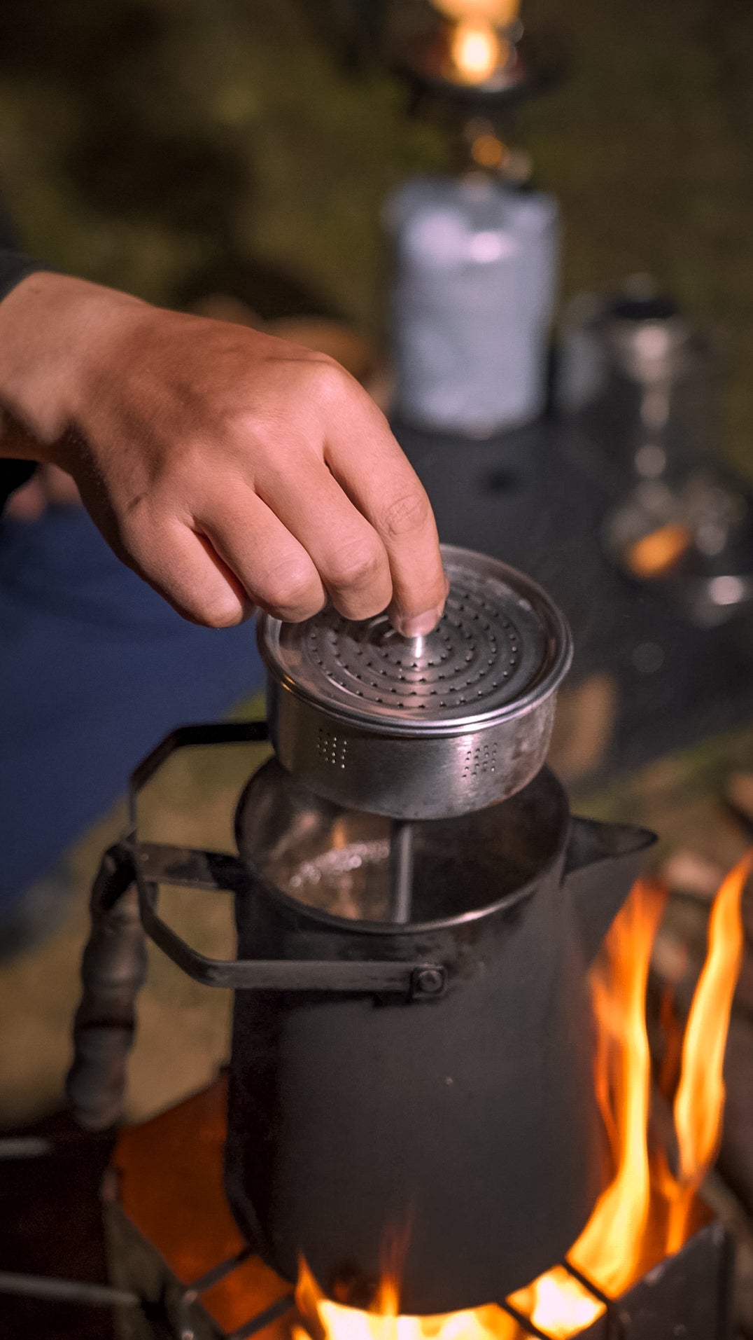 Person using a Nomadia CampStation Stainless Steel Coffee Pot strainer over a camping stove with flames - camping with a fire - outdoor coffee set - outdoor cooking equipment