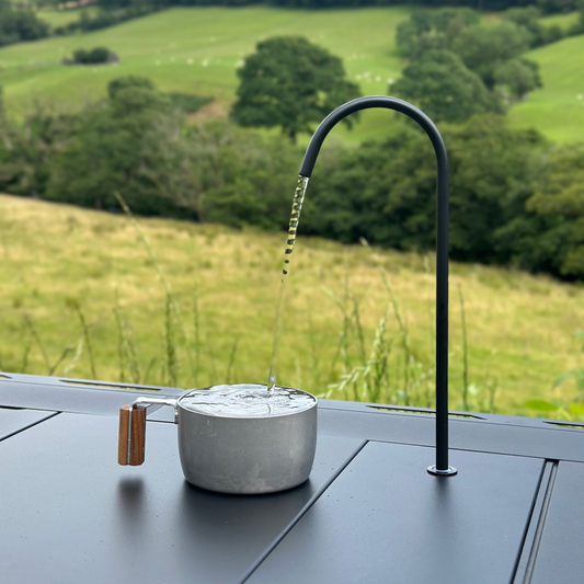 using Nomadia CampStation Water Dispenser to fill Metal pot with water against a scenic background of green fields and trees - camping gear - glamping