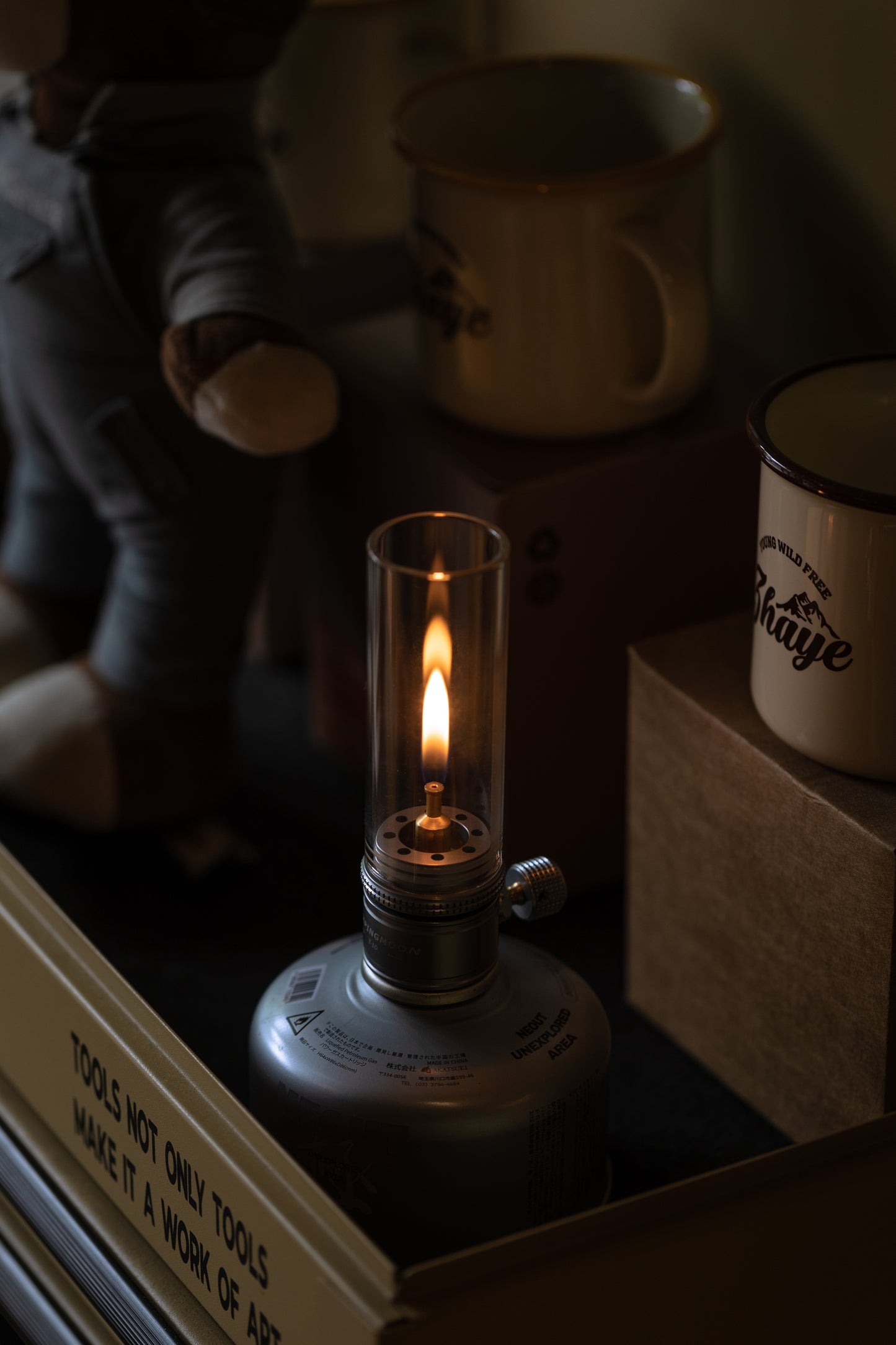 Nomadia Camping Light Atmospheric Camping gas Lamp with a lit flame on a surface with books and mugs in the background - glamping - uk camping