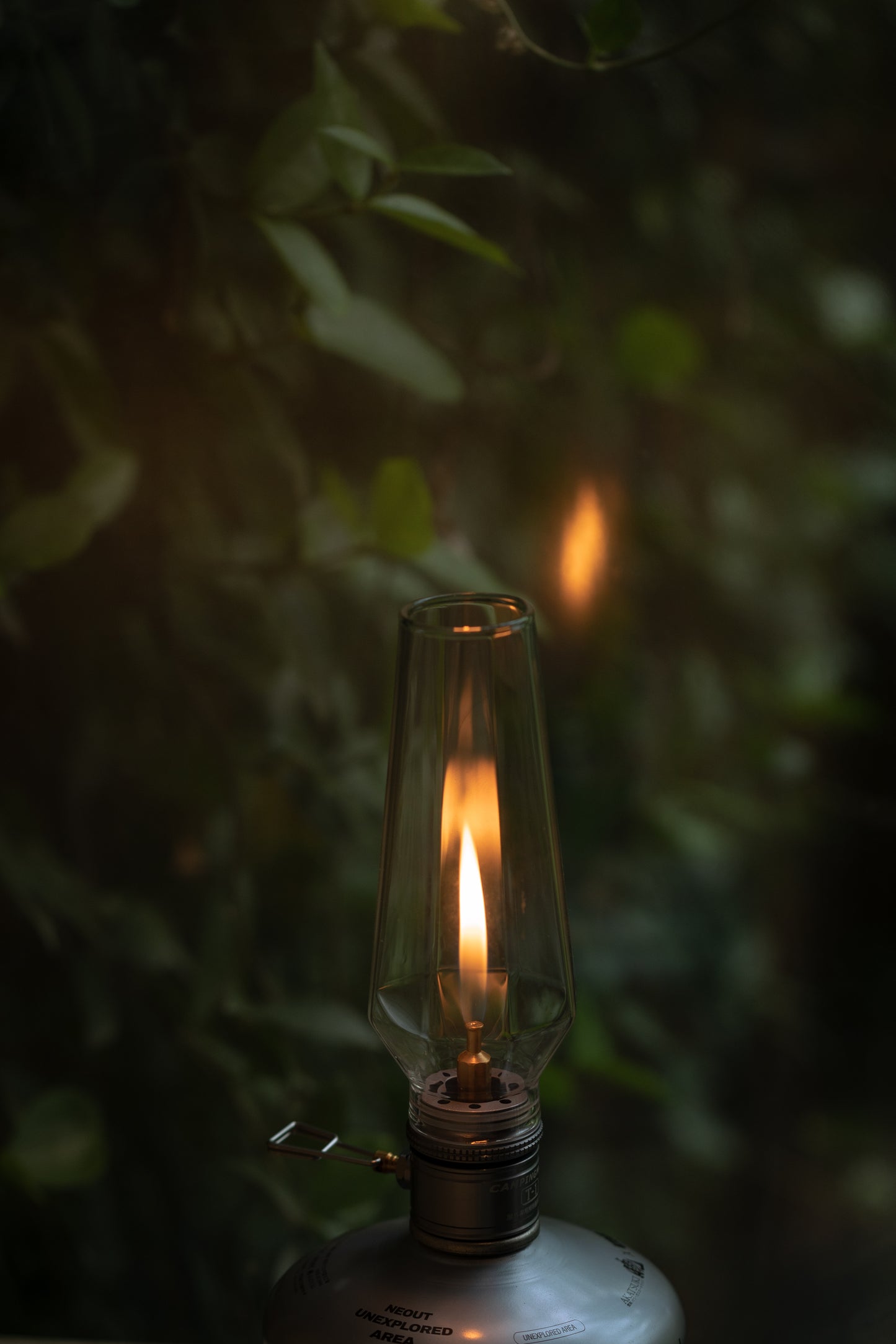 Vintage-style gas lamp with a lit wick against a dark, leafy background - wildcamping accessory - camping accessory - camping with a fire
