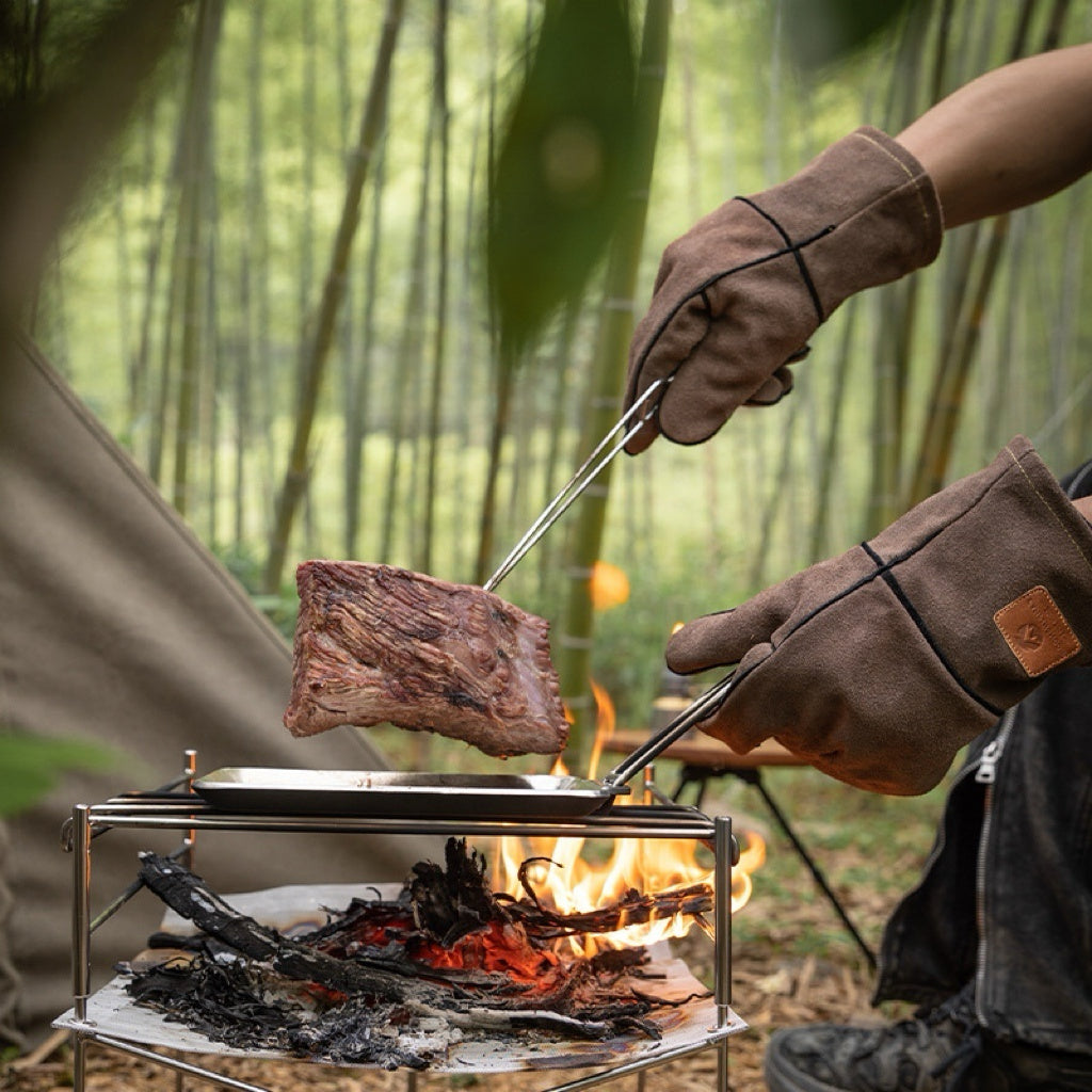 Person grilling meat over a campfire with Nomadia Flamo Heavy-Duty Leather Fire Gloves - camping with a fire - camping gear - muti-use -bbq grill accessory