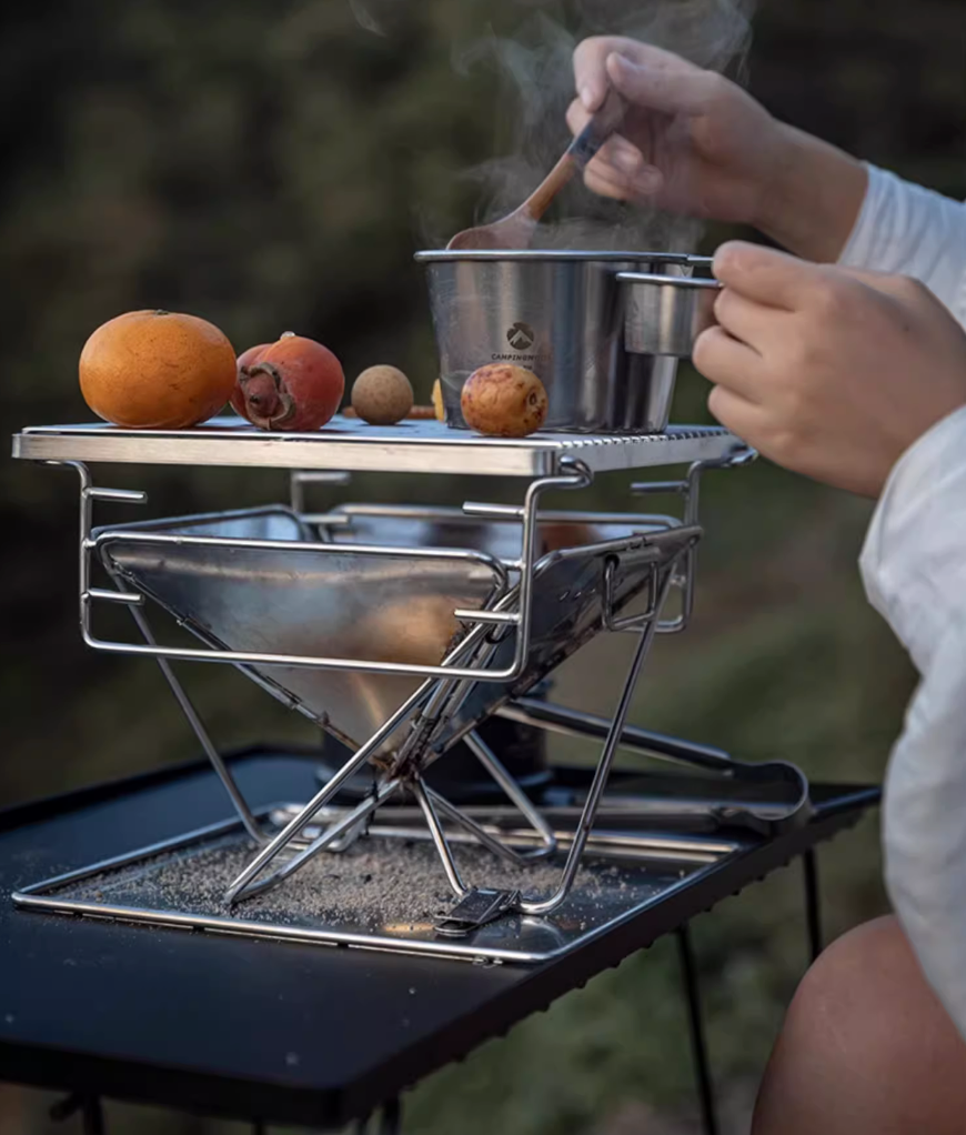 Person cooking outdoors using Nomadia's Flamo Portable Folding BBQ Grill and Fire Pit mini size with fruits and vegetables. - camping gear - uk camping