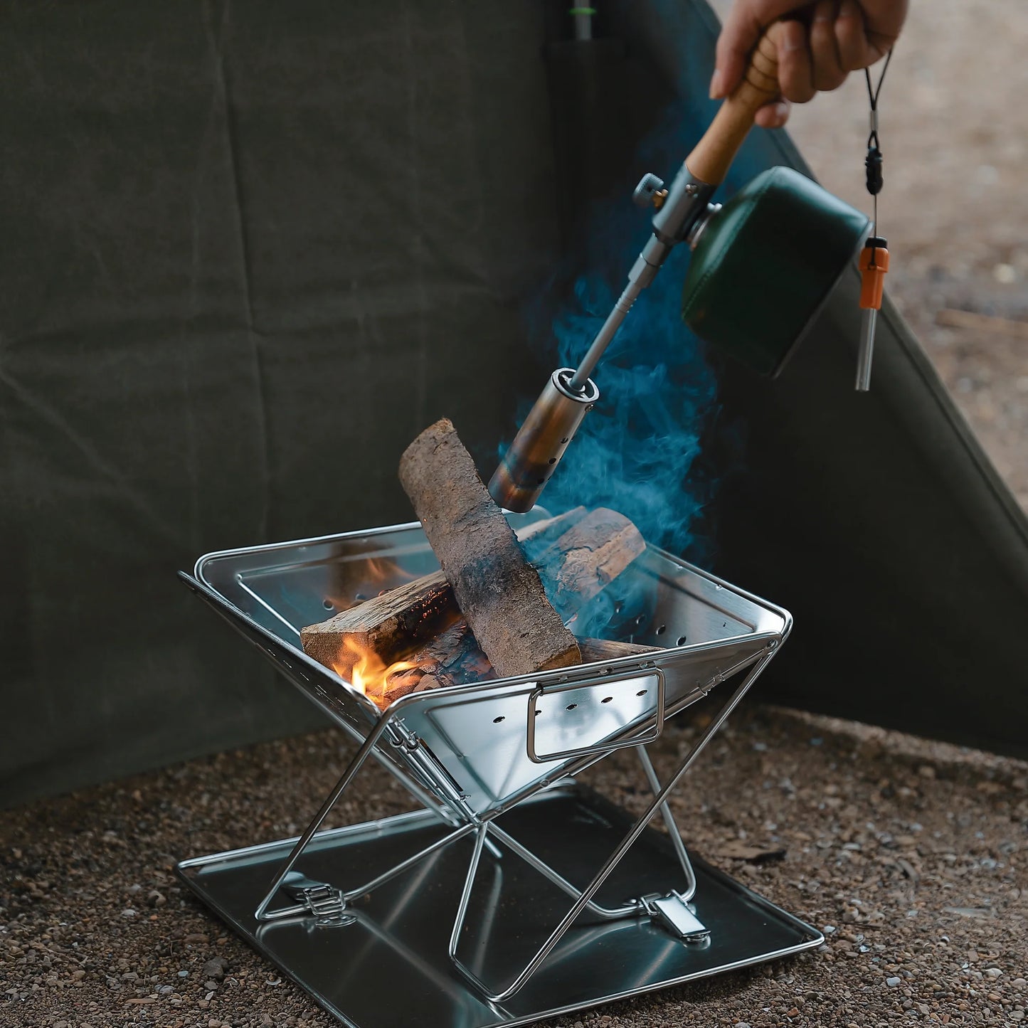 Person using a handheld device to ignite a small fire pit with wood on Nomadia's Flamo Portable Folding BBQ Grill and Fire Pit - wildcamping - camping with a fire - uk camping