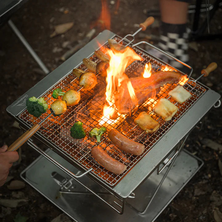 Outdoor camping scene with Nomadia's Flamo Portable Folding BBQ Grill and Fire Pit over an open flame, cooking sausages and vegetables - wildcamping - camping with a fire - uk camping