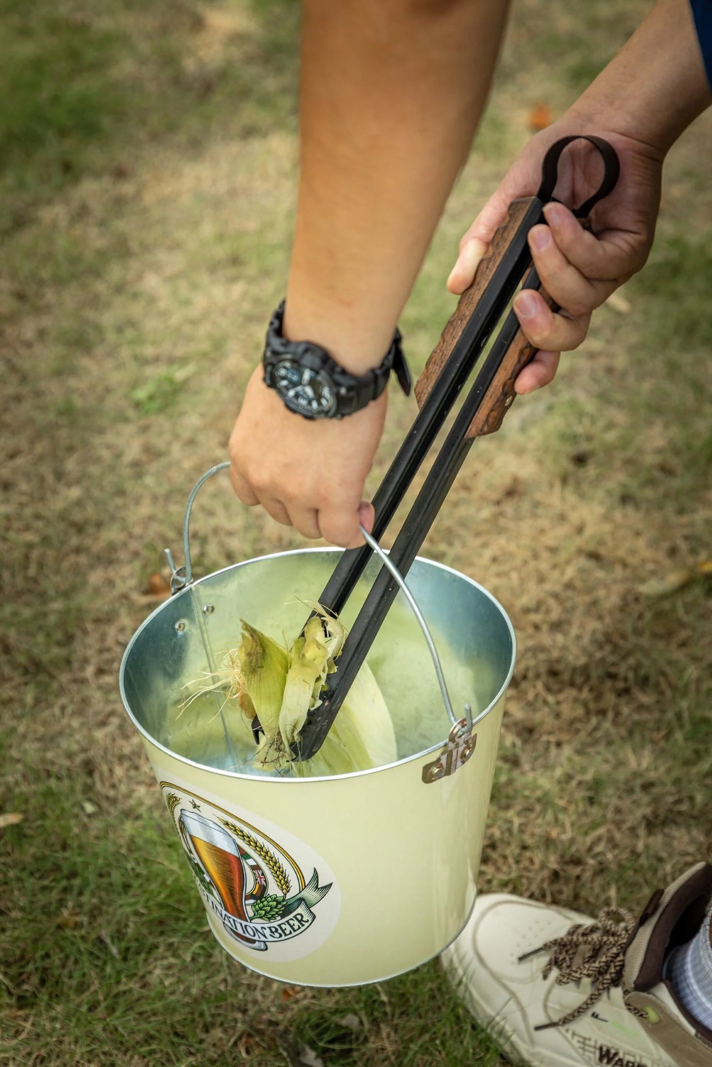 person using Nomadia Flamo Wood Handle Outdoor Tongs to pick up trash and put into a basket - camping gear - bbq grill accessory - camping essential