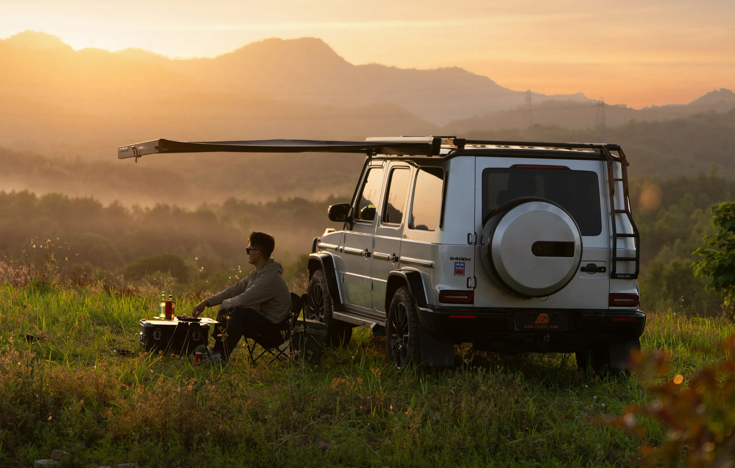 Man sitting by a parked white SUV in a scenic landscape White SUV with Nomadia Glide World's fastest opening awning aluminium awning - outdoor tent - quick setup tent - easy setup - all weather ready - wildcamping - overlanding