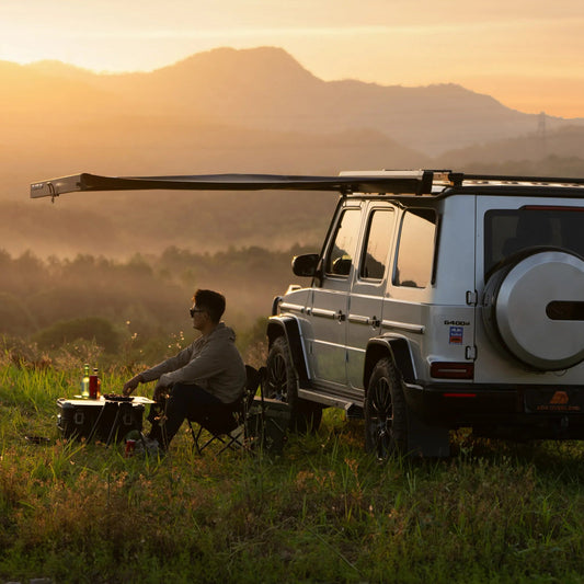 Man sitting by a parked white SUV in a scenic landscape White SUV with Nomadia Glide World's fastest opening awning aluminium awning - outdoor tent - quick setup tent - easy setup - all weather ready - wildcamping - overlanding