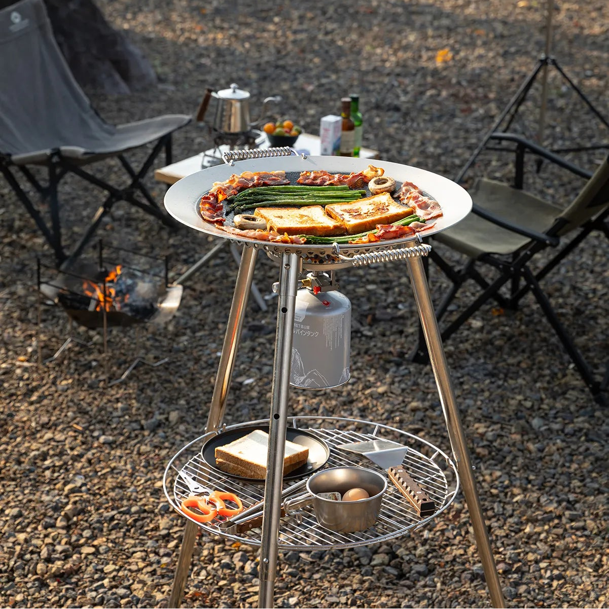 Outdoor camping cooking scene with Nomadia Horizon Cook portable outdoor cooking station for sharing and plateful of food on a gravel ground - portable grill pan - outdoor cookware for groups - multi-function camping stove - vanlife cooking gear - campervan kitchen setup