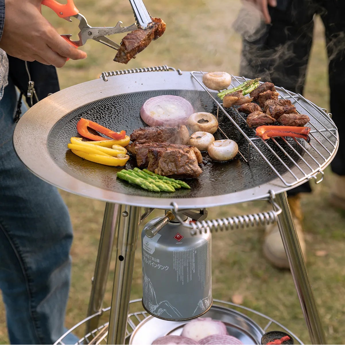Portable grill with food being cooked outdoors using Nomadia Horizon Cook portable outdoor cooking station for sharing, with a person using tongs to handle the food - outdoor cookware for groups - multi-function camping stove