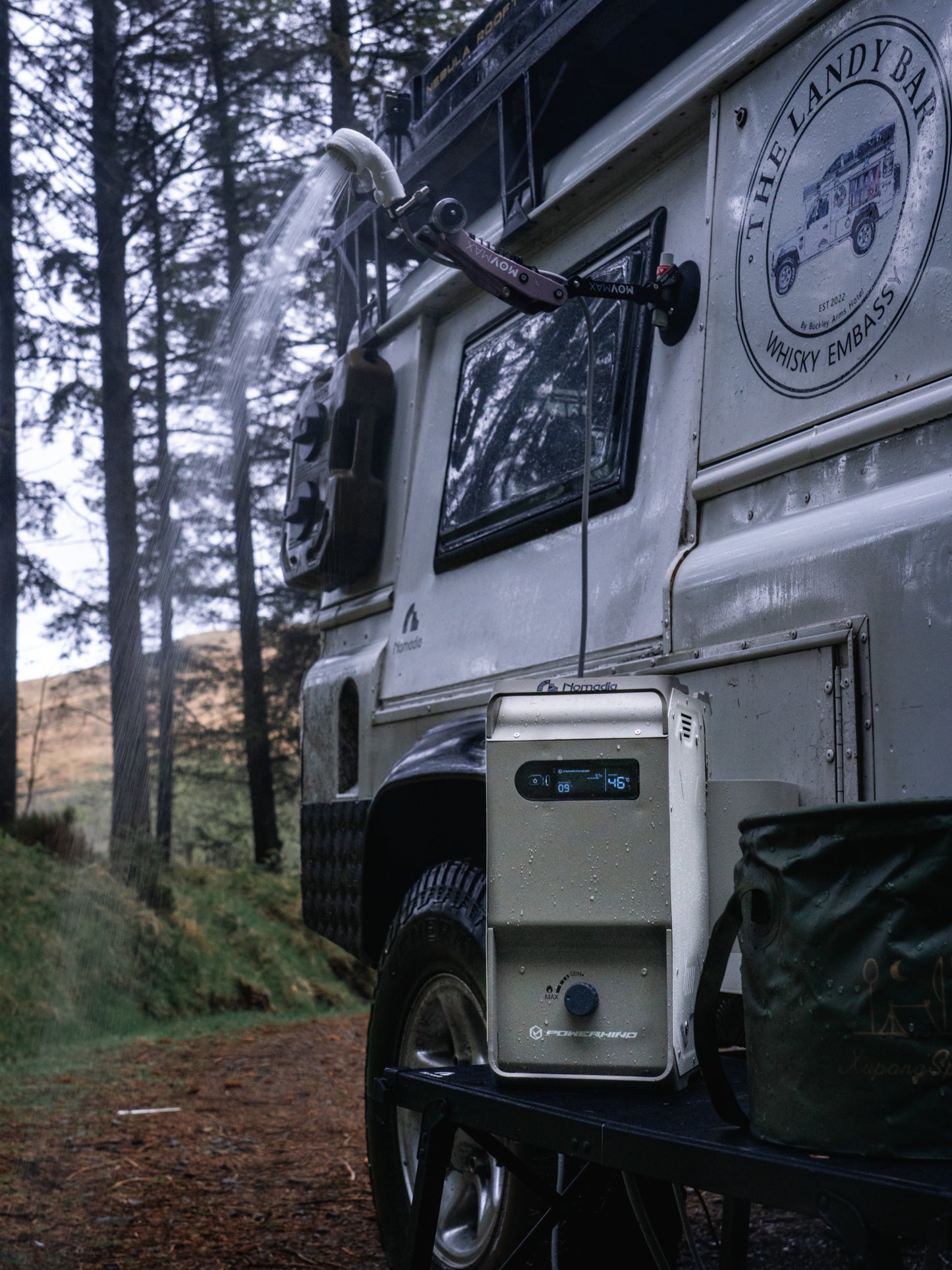 Nomadia ThermoFlow Portable Heated Shower next to a white van with a water hose attached to the van and hot water running in a forest. wildcamping - camping gear - uk camping