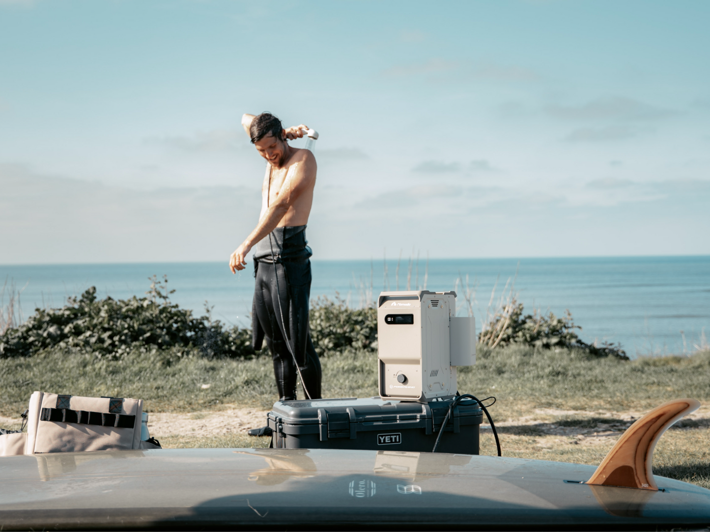 a male surfer standing on a grassy area by the ocean taking shower using thermoflow - a portable heated shower system from nomadia - open air - camping gear - uk camping - outdoorsy