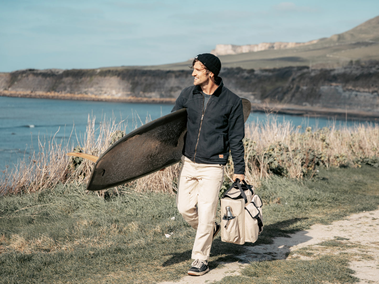a male surfer walking along a coastal path holding a surfboard and carrying a bag with nomadia's protable heated shower system ThermoFlow inside - outdoorsy - camping gear - uk camping