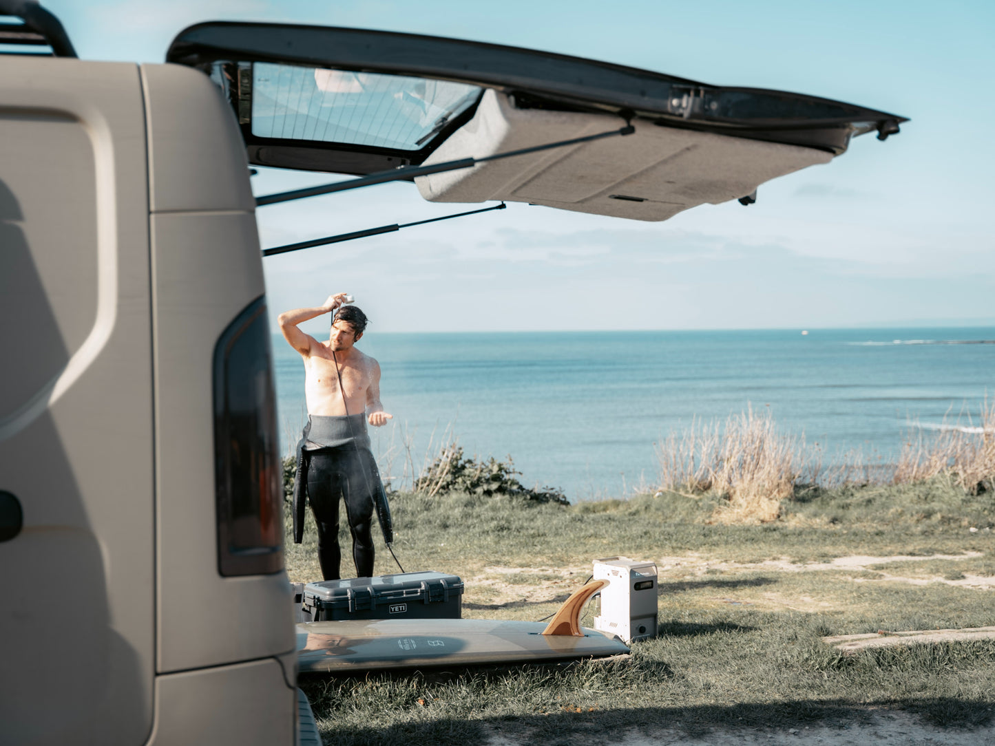 Man standing next to a van with an open door by the ocean, taking shower using Nomadia's ThermoFlow Portable Heated Shower for Camping and outdoor use