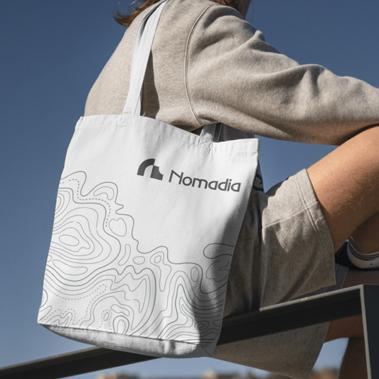 Person holding a white tote bag with 'Nomadia' logo against a clear blue sky. canva bag - print bag - bio-friendly