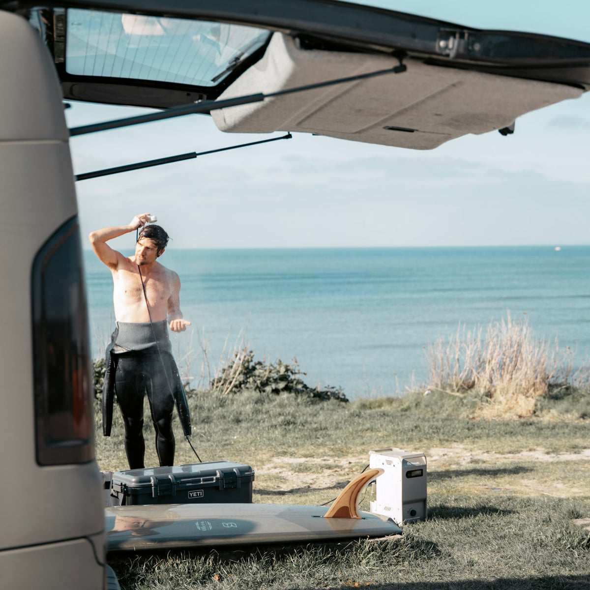 A person showering with Nomadia ThermoFlow beside a campervan and surfboard overlooking the sea.