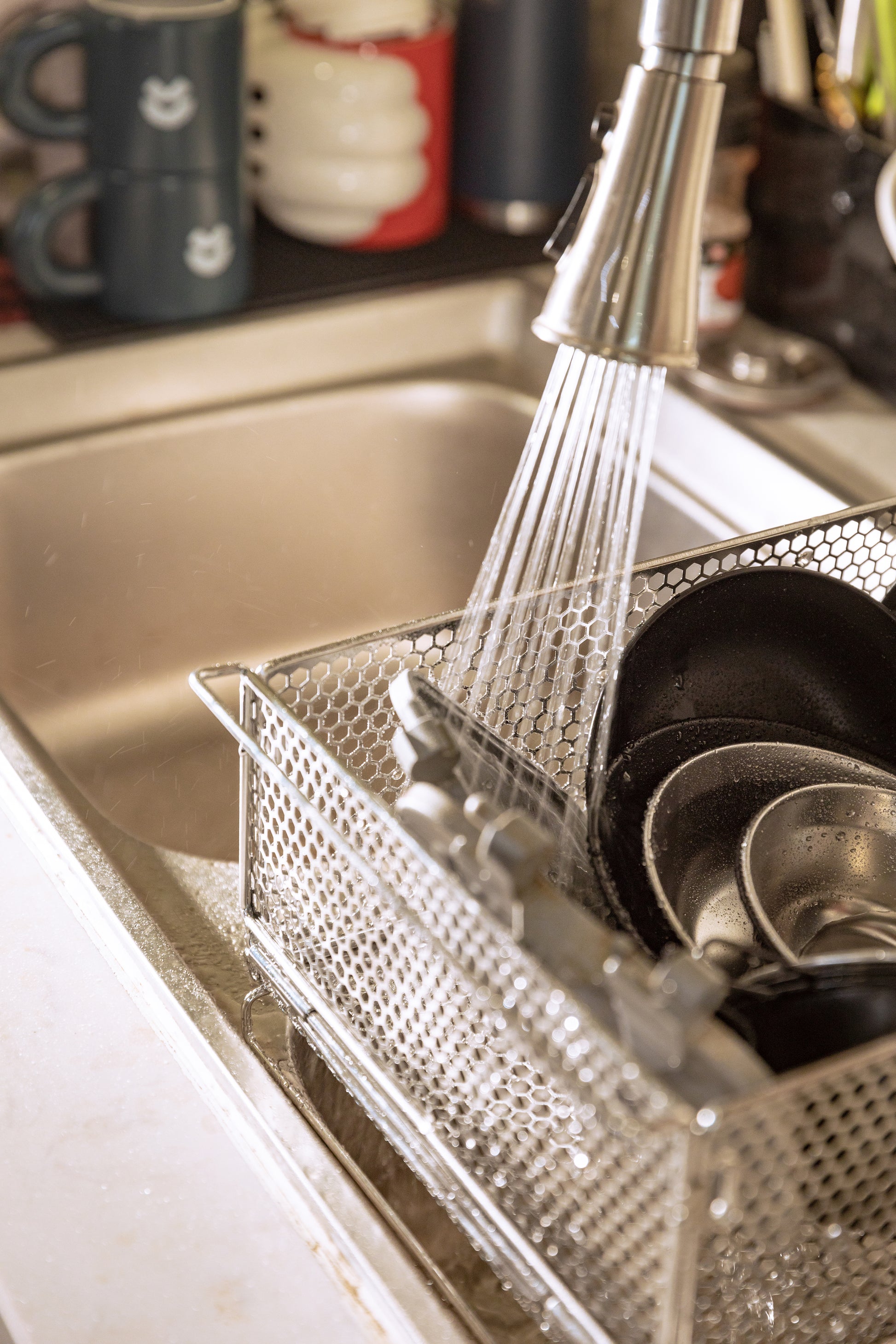 metal mesh basket used as dish rack with dishes being rinsed under a running faucet in a kitchen sink - multi-use camping gear - multi-purpose