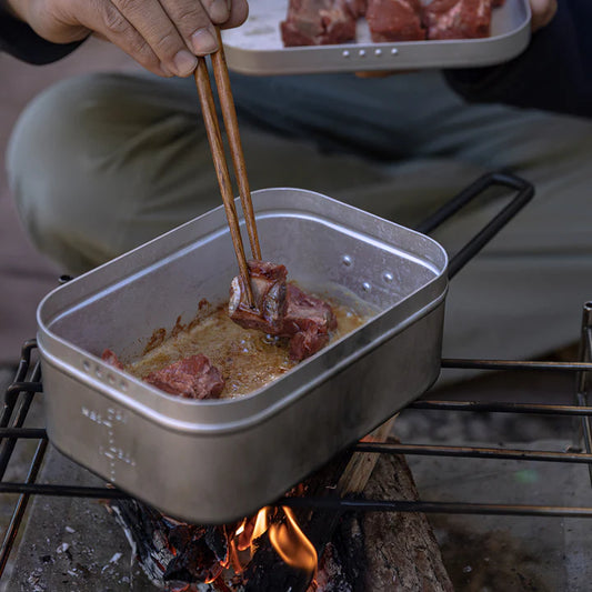 Person cooking meat over a fire using Nomadia CampStation Outdoor Aluminium Lunch Box and chopsticks - outdoor cooking - cooking utensil - camping essential
