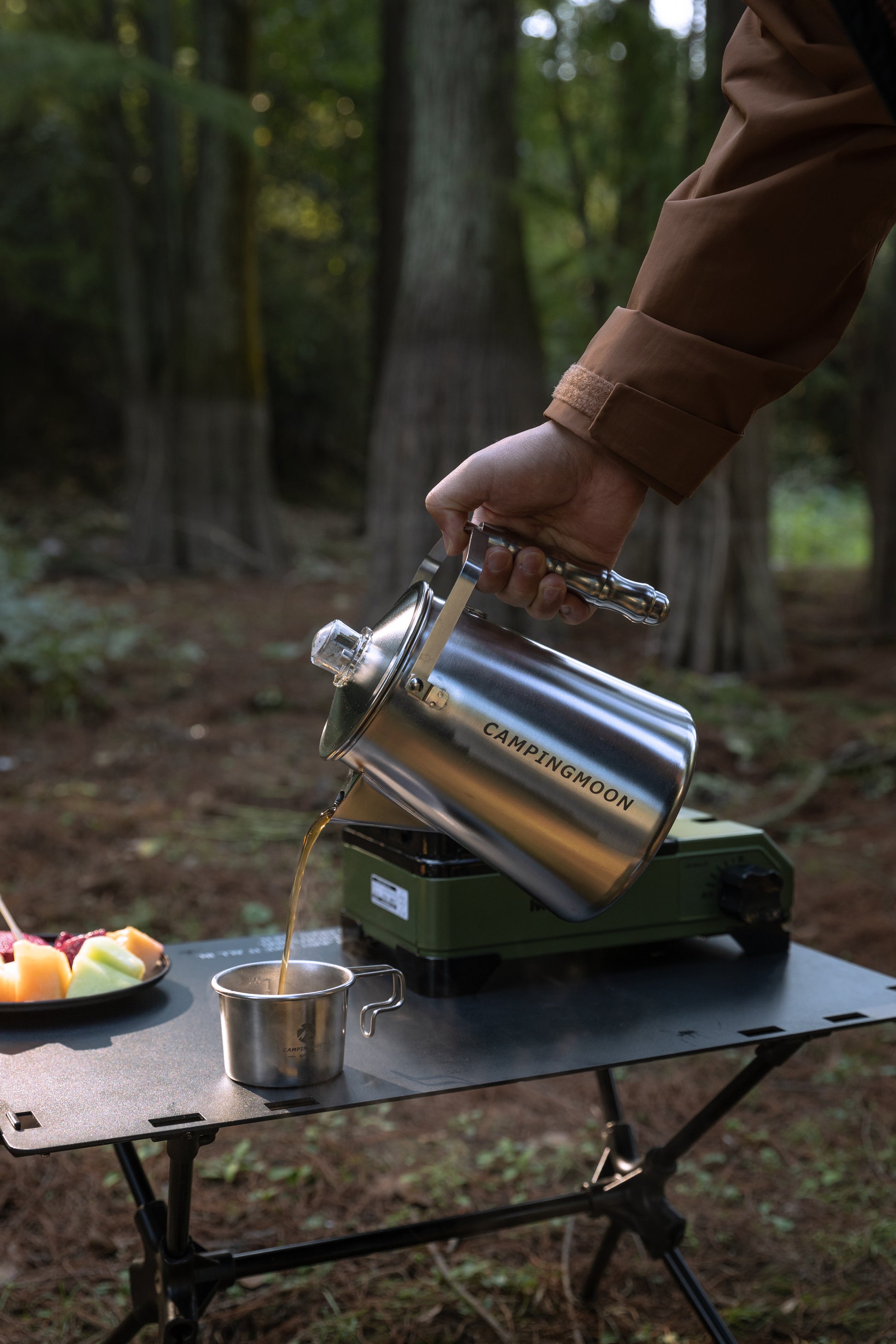 Person pouring hot coffee from a Nomadia CampStation Stainless Steel Coffee Pot into a cup in a forest setting - glamping in uk - outdoor coffee pot - outdoor cooking equipment