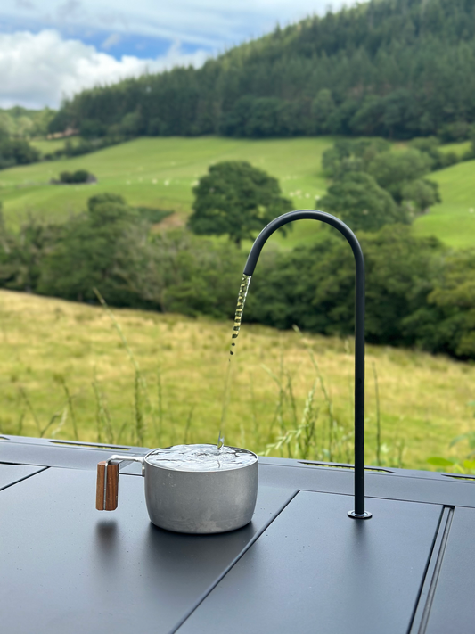 using Nomadia CampStation Water Dispenser to fill Metal pot with water against a scenic background of green fields and trees - camping gear - glamping 