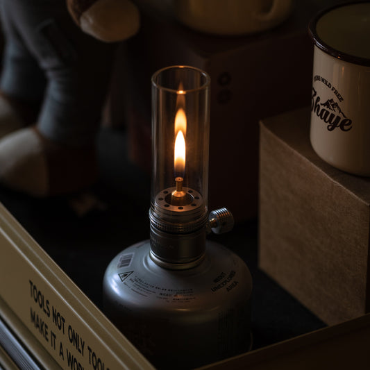 Nomadia Camping Light Atmospheric Camping gas Lamp with a lit flame on a surface with books and mugs in the background - glamping - uk camping