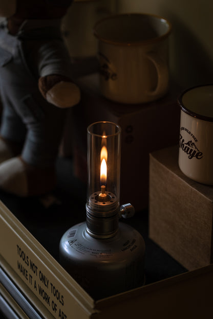 Nomadia Camping Light Atmospheric Camping gas Lamp with a lit flame on a surface with books and mugs in the background - glamping - uk camping