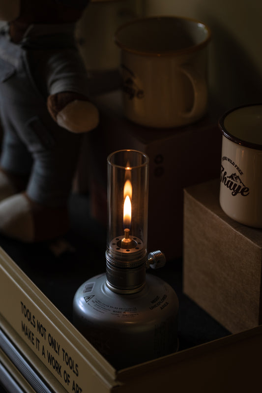 Nomadia Camping Light Atmospheric Camping gas Lamp with a lit flame on a surface with books and mugs in the background - glamping - uk camping