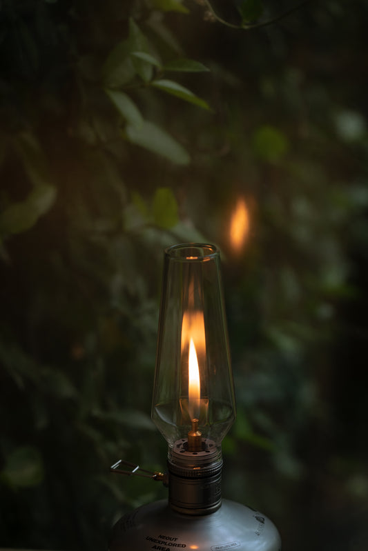 Vintage-style gas lamp with a lit wick against a dark, leafy background - wildcamping accessory - camping accessory - camping with a fire