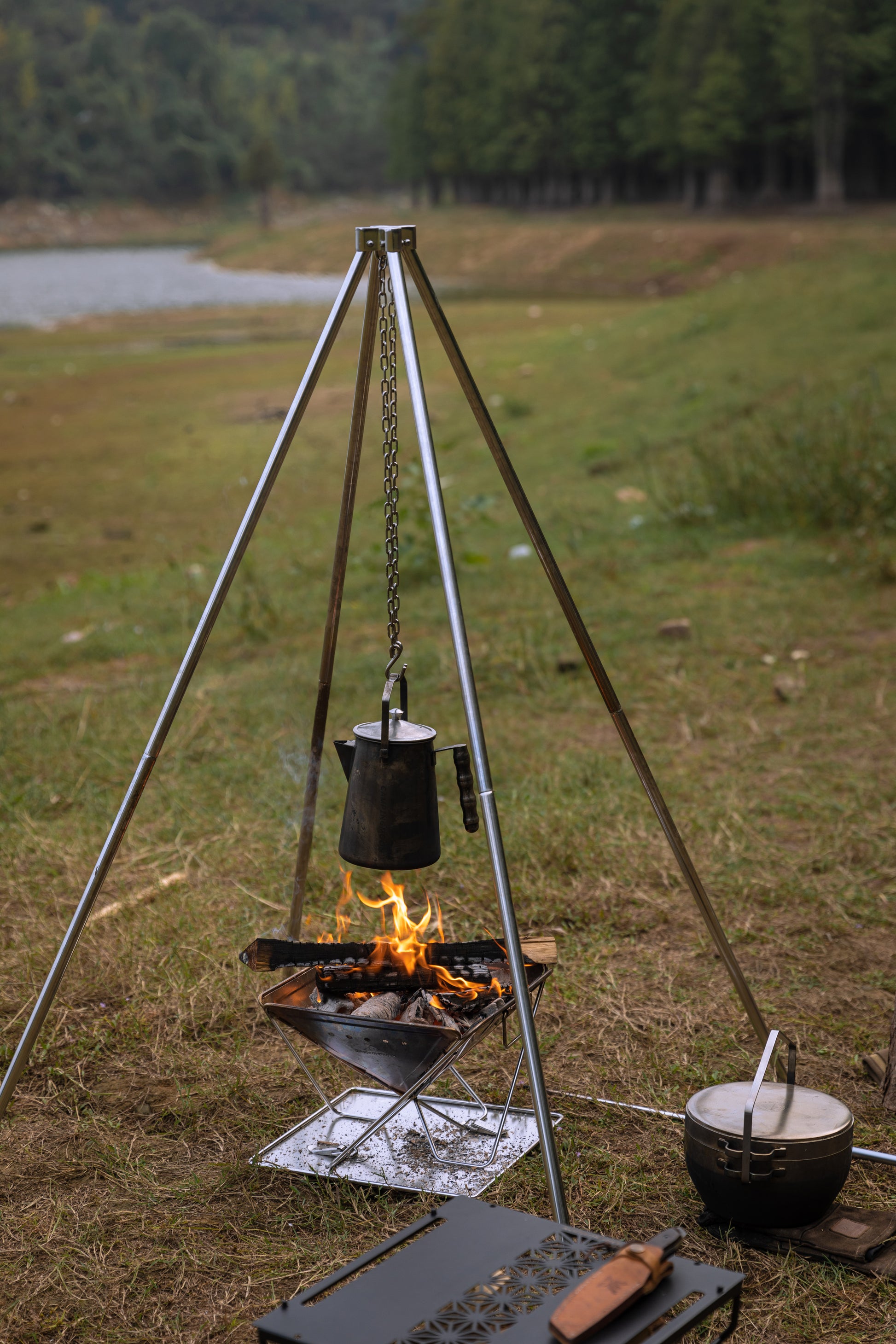 Outdoor camping setup with tripod quadripod fire pit and Nomadia Flamo Crafted Camping Kettle over a fire pit in a natural setting. camping with fire - wild camping