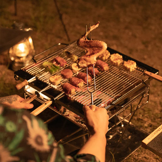 Person grilling skewered meat and vegetables on Nomadia's Flamo Portable Folding BBQ Grill and Fire Pit at night - wildcamping - uk camping