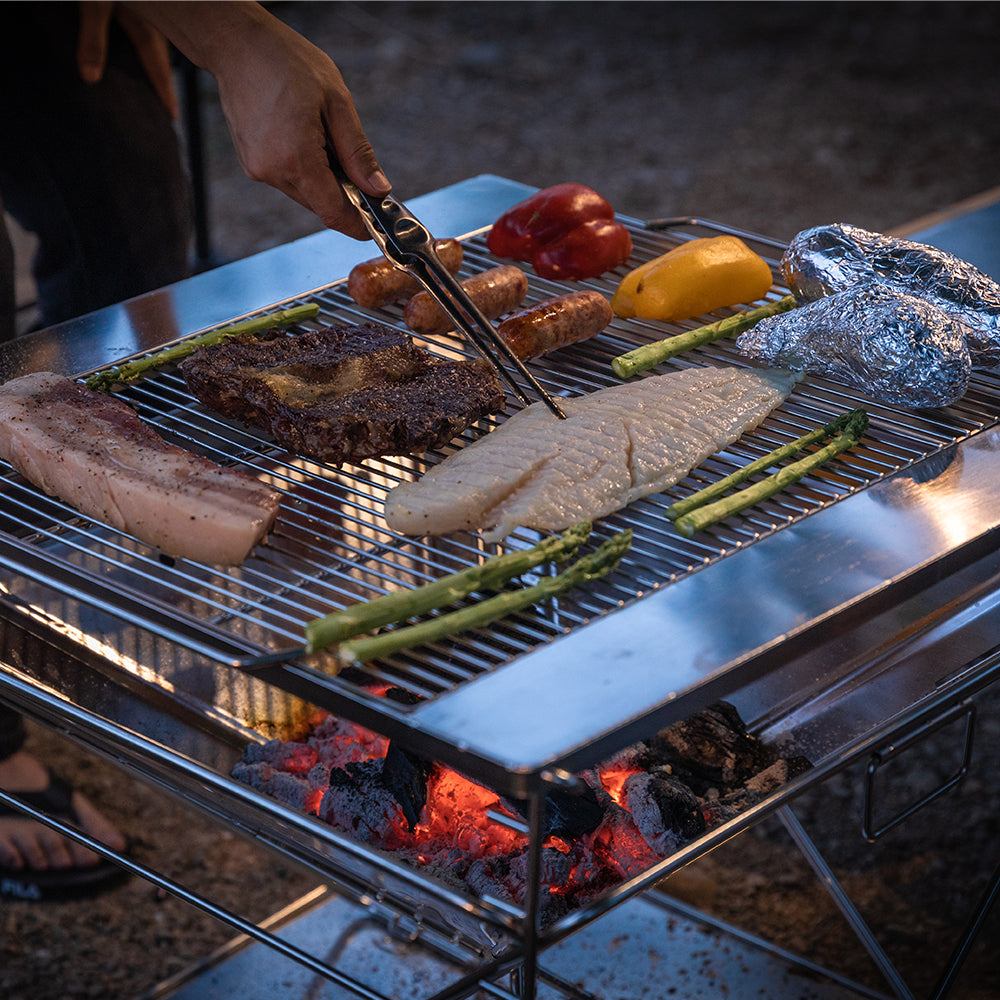 Grilling meat, vegetables, and sausages on Nomadia's Flamo Portable Folding BBQ Grill and Fire Pit with food being cooked on a natural outdoor setting with a person using tongs.- wildcamping - camping with a fire - uk camping 