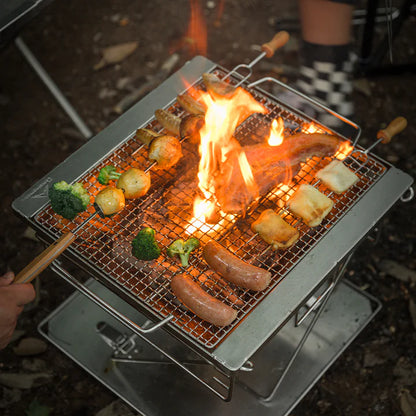 Outdoor camping scene with Nomadia's Flamo Portable Folding BBQ Grill and Fire Pit over an open flame, cooking sausages and vegetables - wildcamping - camping with a fire - uk camping