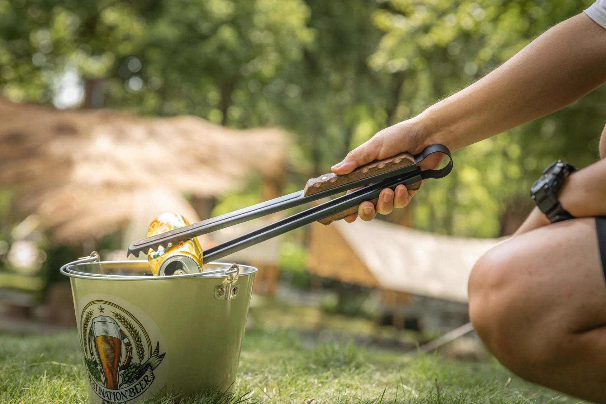 Person using Nomadia Flamo Wood Handle Outdoor Tongs to pick up trash in a metal container outdoors with trees in the background - camping gear - camping accessory- uk camping