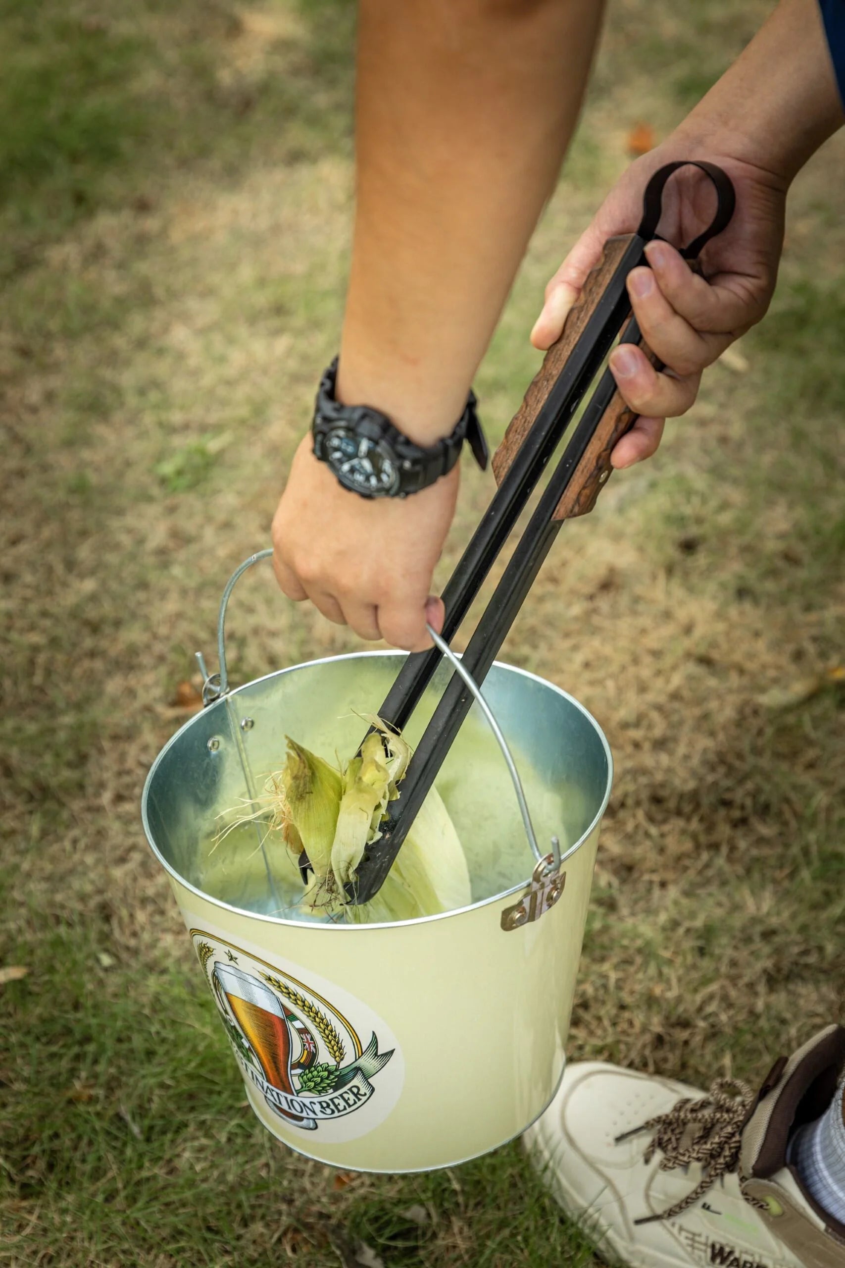 person using Nomadia Flamo Wood Handle Outdoor Tongs to pick up trash and put into a basket - camping gear - bbq grill accessory - camping essential