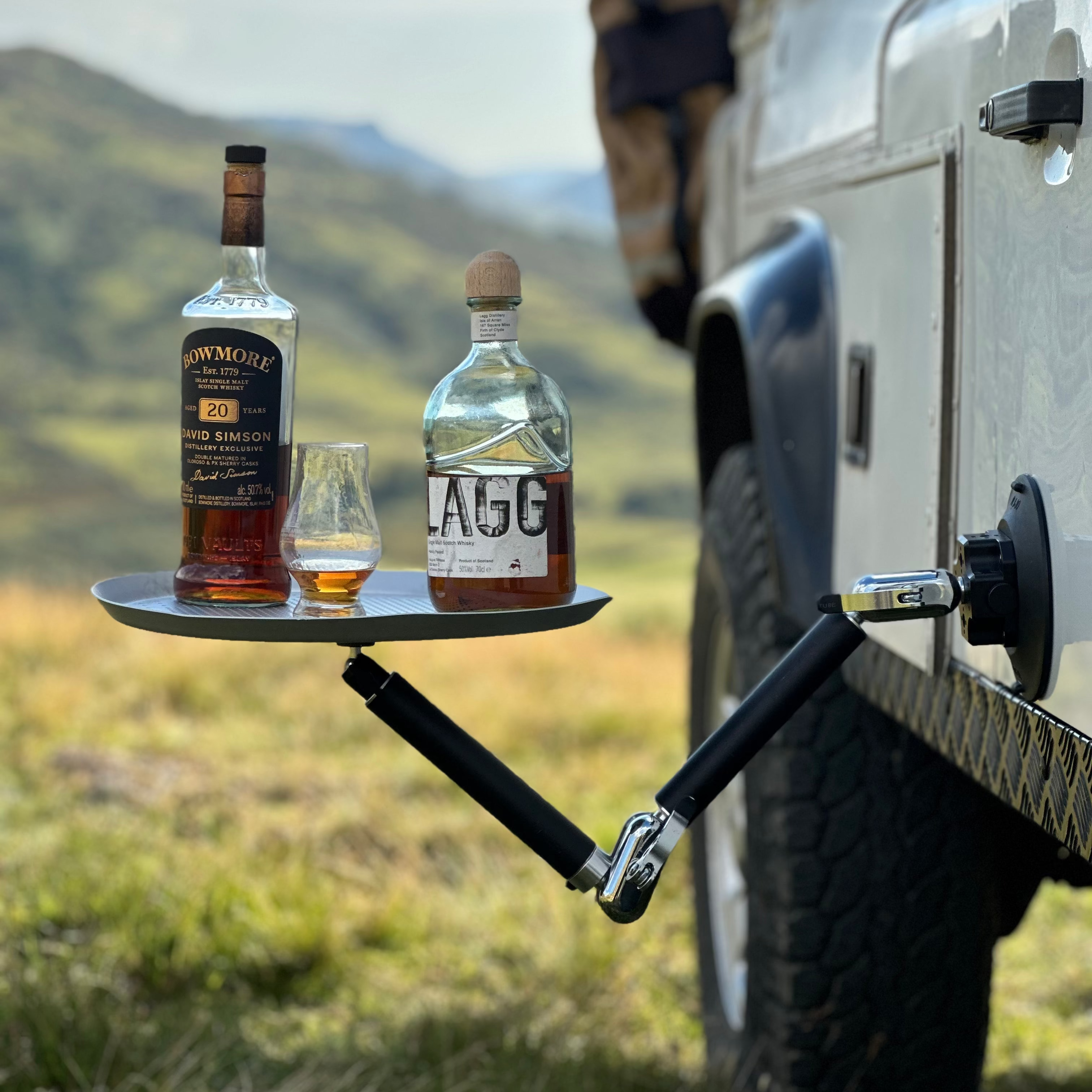 Two bottles of alcohol and a glass on a tray attached to a vehicle using Nomadia FlexMount Suction Arm with mountains in the background - camping and survival - uk camping - strong powerful and reliable