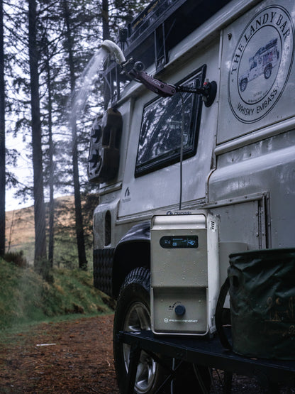 Nomadia ThermoFlow Portable Heated Shower next to a white van with a water hose attached to the van and hot water running in a forest. wildcamping - camping gear - uk camping
