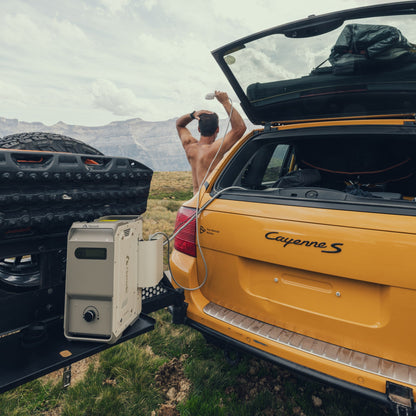 A traveller using the ThermoFlow shower beside an off-road vehicle in a mountain landscape.